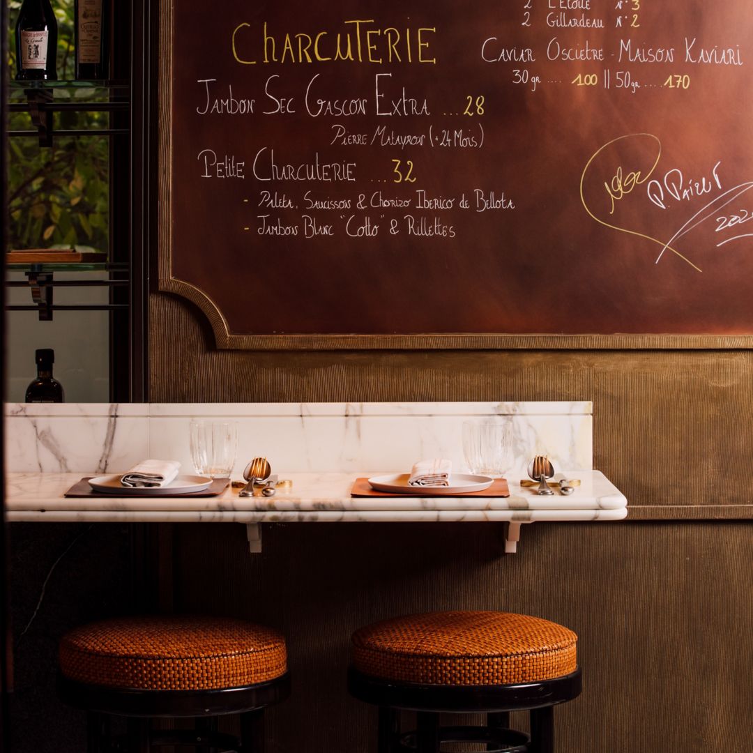 A chalkboard menu listing various types of charcuterie and cheese, with a counter and stools in the foreground.